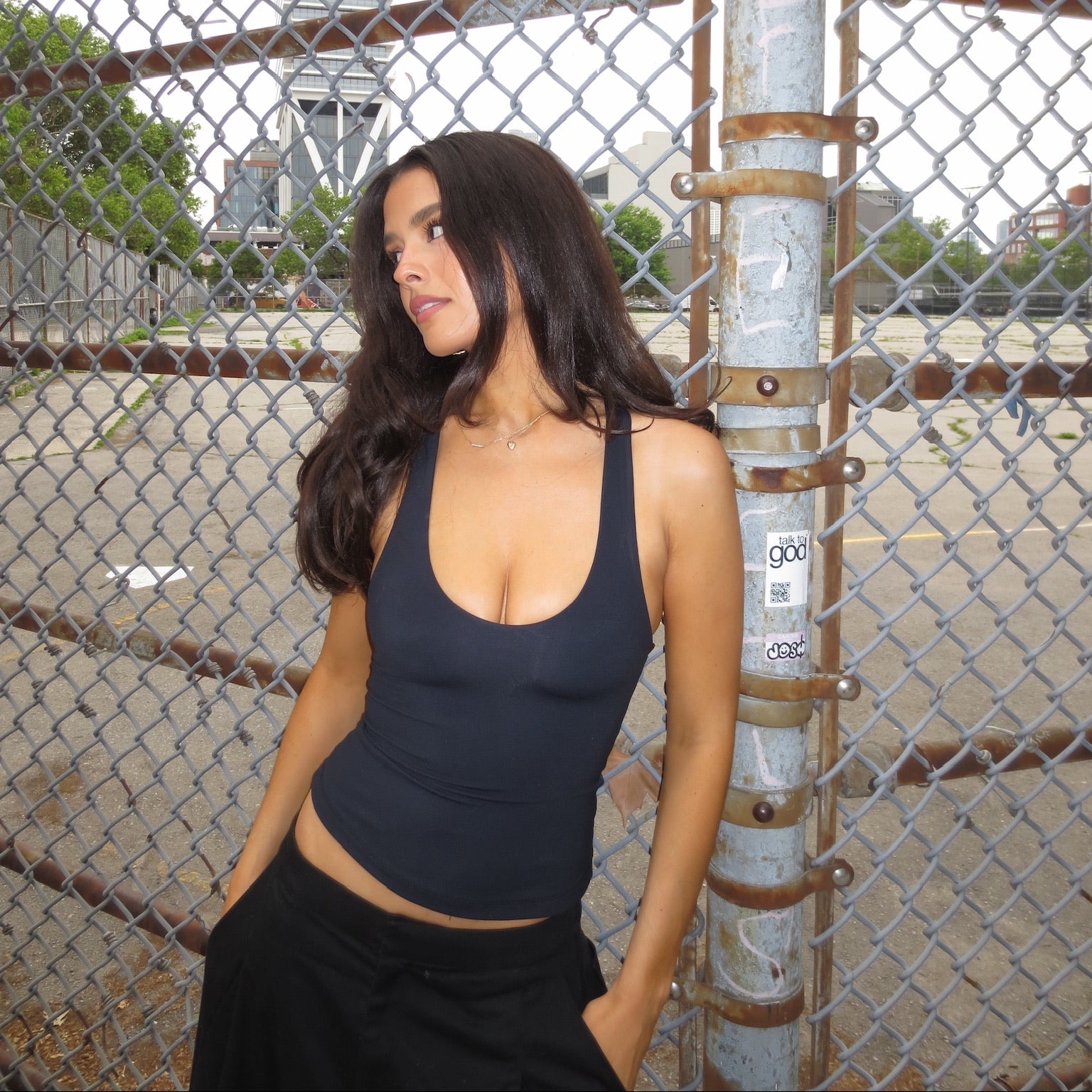 A woman with long dark hair wearing a black scoop-neck tank top and black pants, posing in front of a chain-link fence with a city skyline in the background.