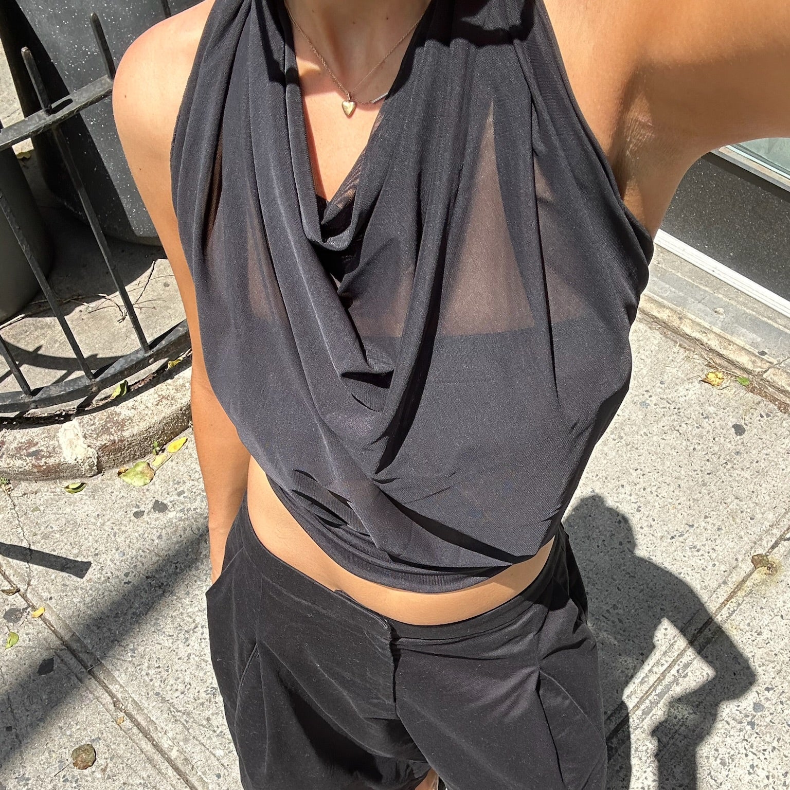 A person outdoors on a sidewalk wearing a black draped halter-style top and black wide-leg pants, with black flip-flops. The photo is taken from above at a slight angle in bright daylight.