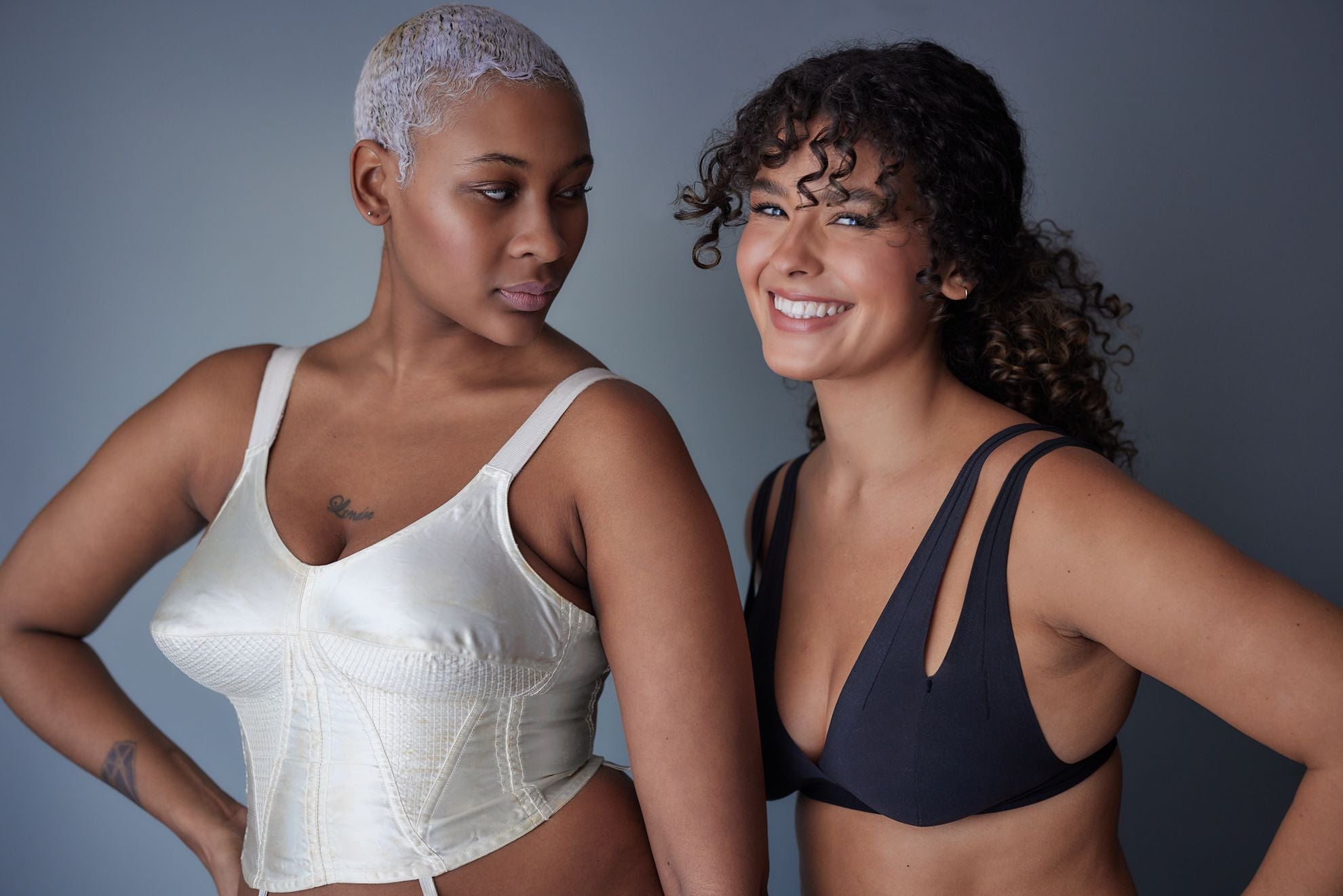 Two women standing, one wearing a corset and the other the Nuudii Tee System in Black