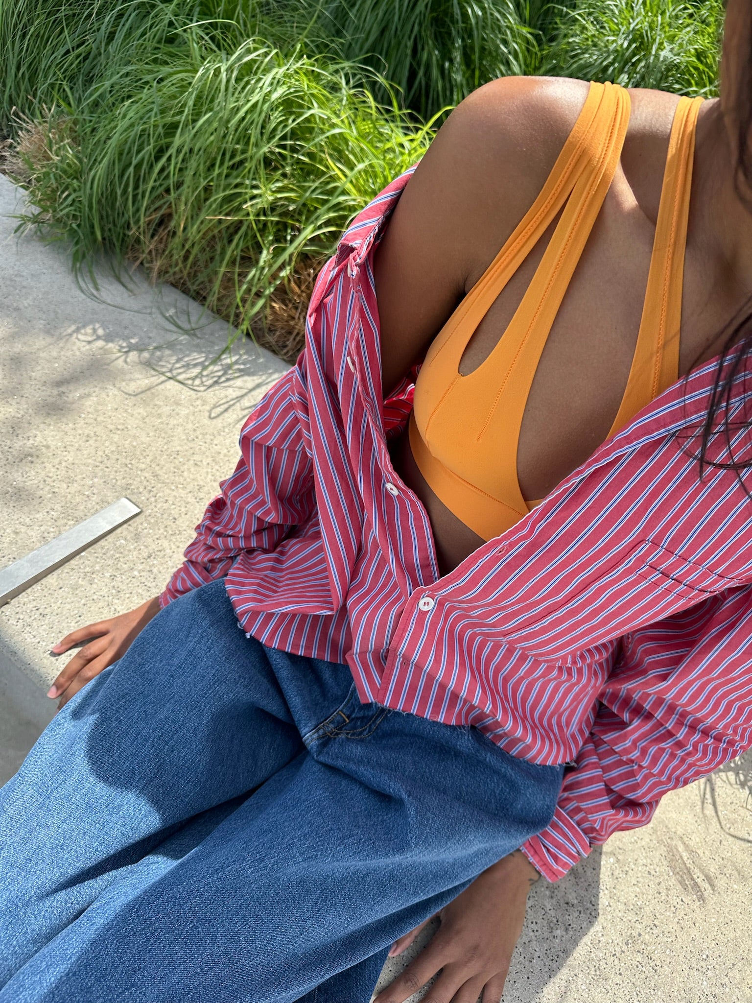 A close-up lifestyle shot of a person wearing a bright orange nuudii top, a pink and white striped button-down shirt, and blue jeans, sitting outdoors on a concrete surface with green plants in the background.