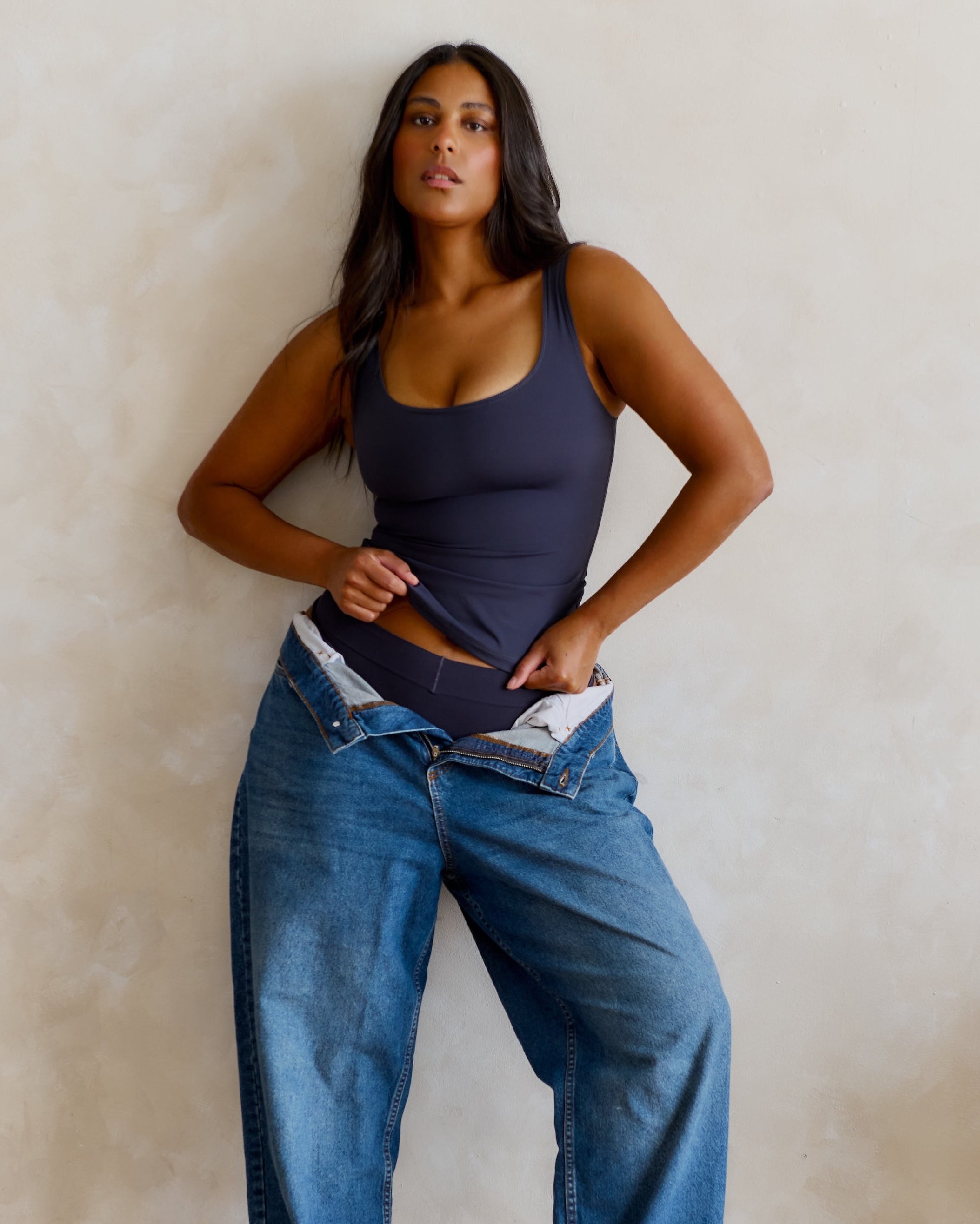 A woman against a textured wall, wearing oversized blue jeans and a myst tank top.