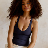 A woman with voluminous curly brown hair wearing a myst racerback top and white pants, posing against a textured beige wall.