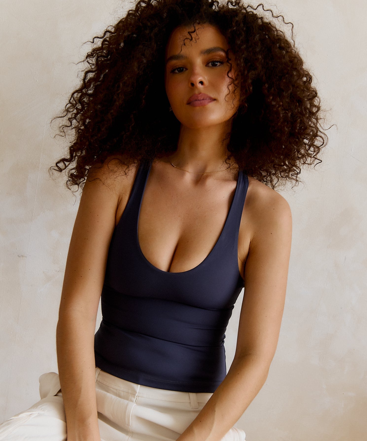 A woman with voluminous curly brown hair wearing a myst racerback top and white pants, posing against a textured beige wall.