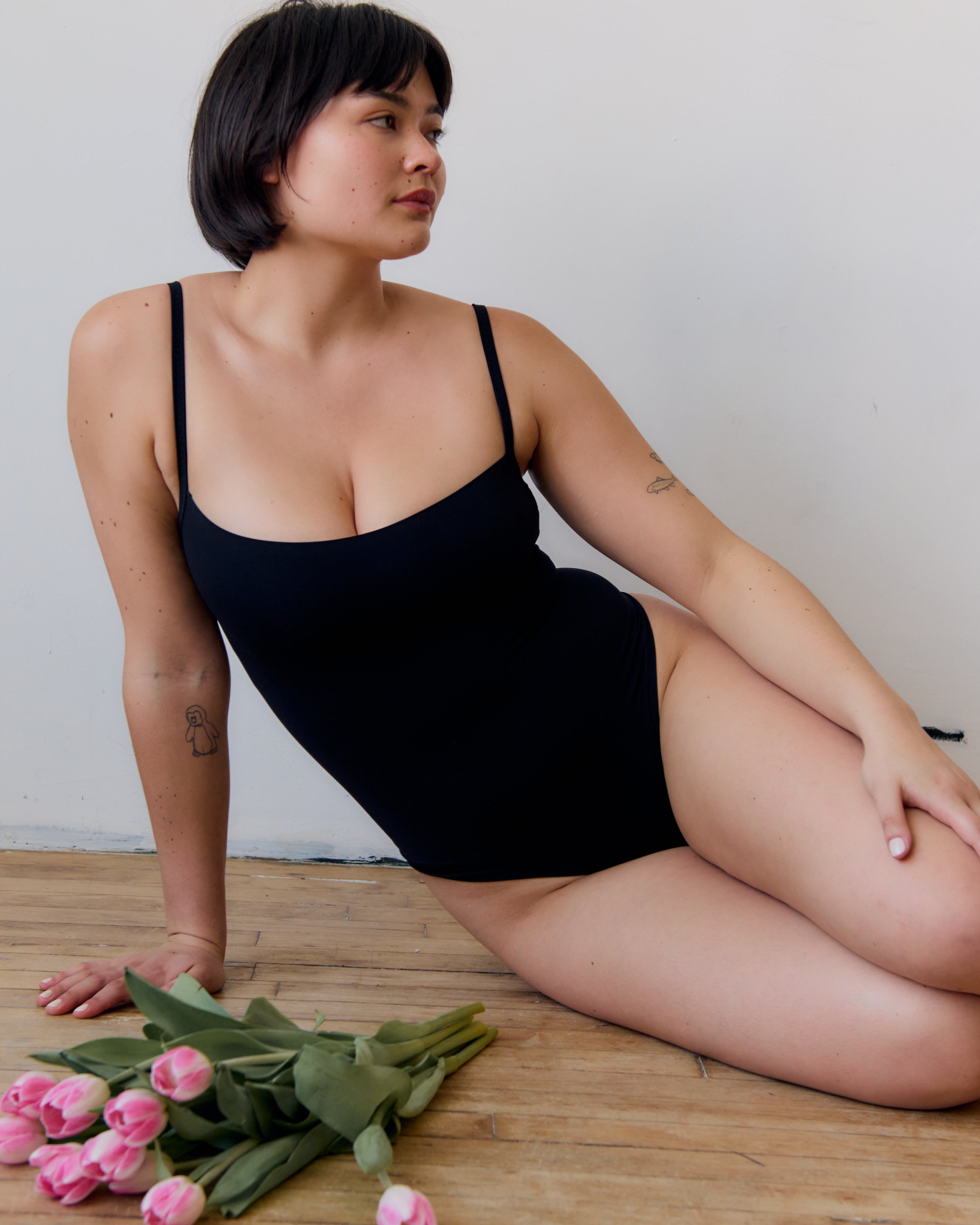 A woman wearing a black bodysuit reclines on a wooden floor against a white wall, with pink tulips in the foreground.