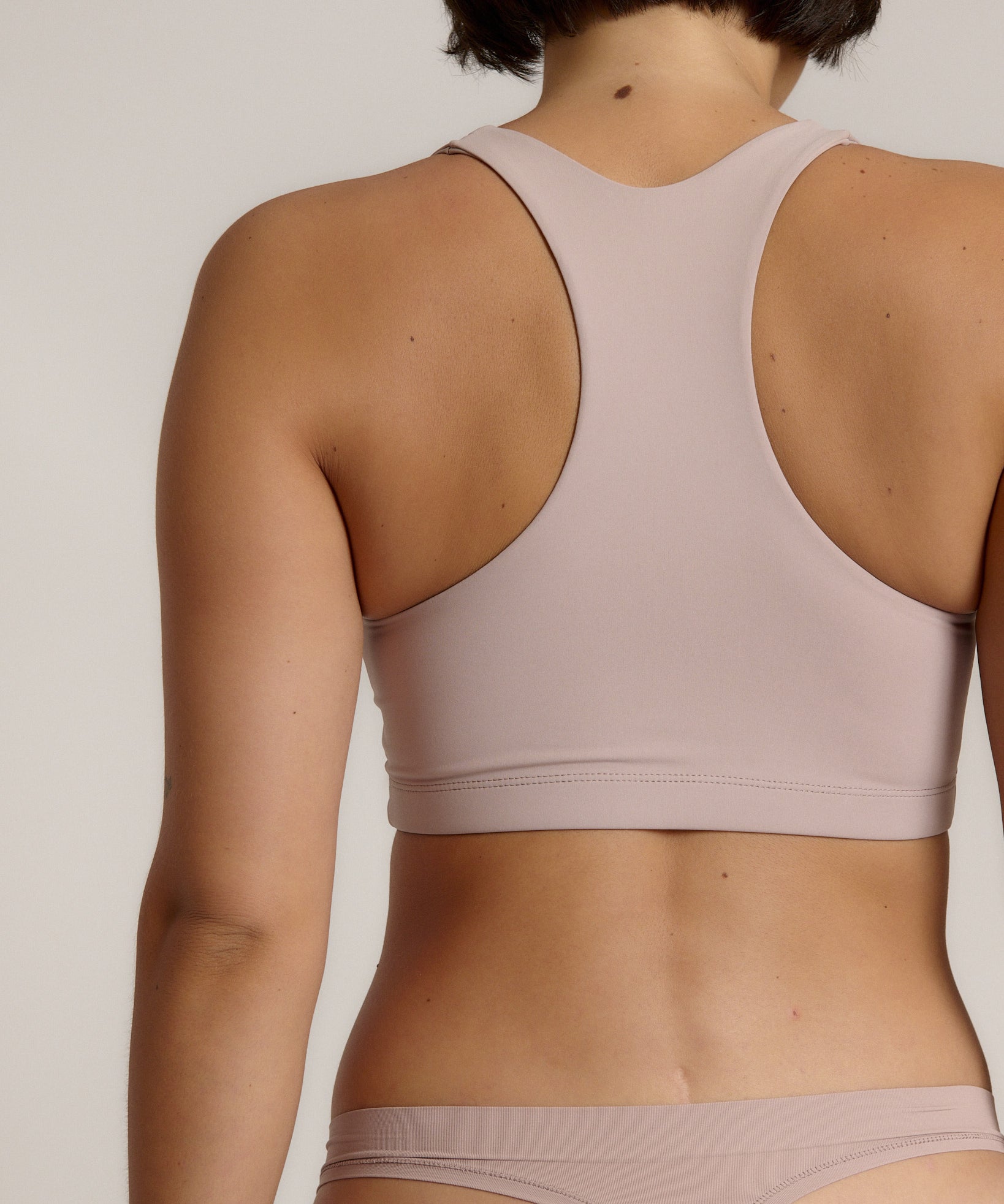 A woman modeling a dusk cropped racerback and matching underwear, against a plain light beige studio background.