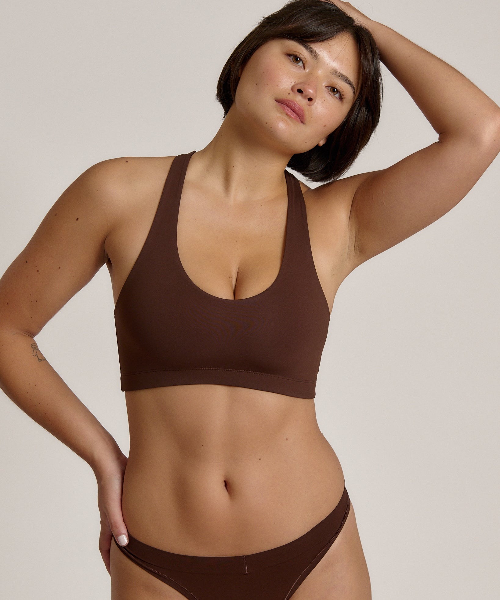 A woman modeling a matching mocha racerback and underwear set against a plain light beige studio background.