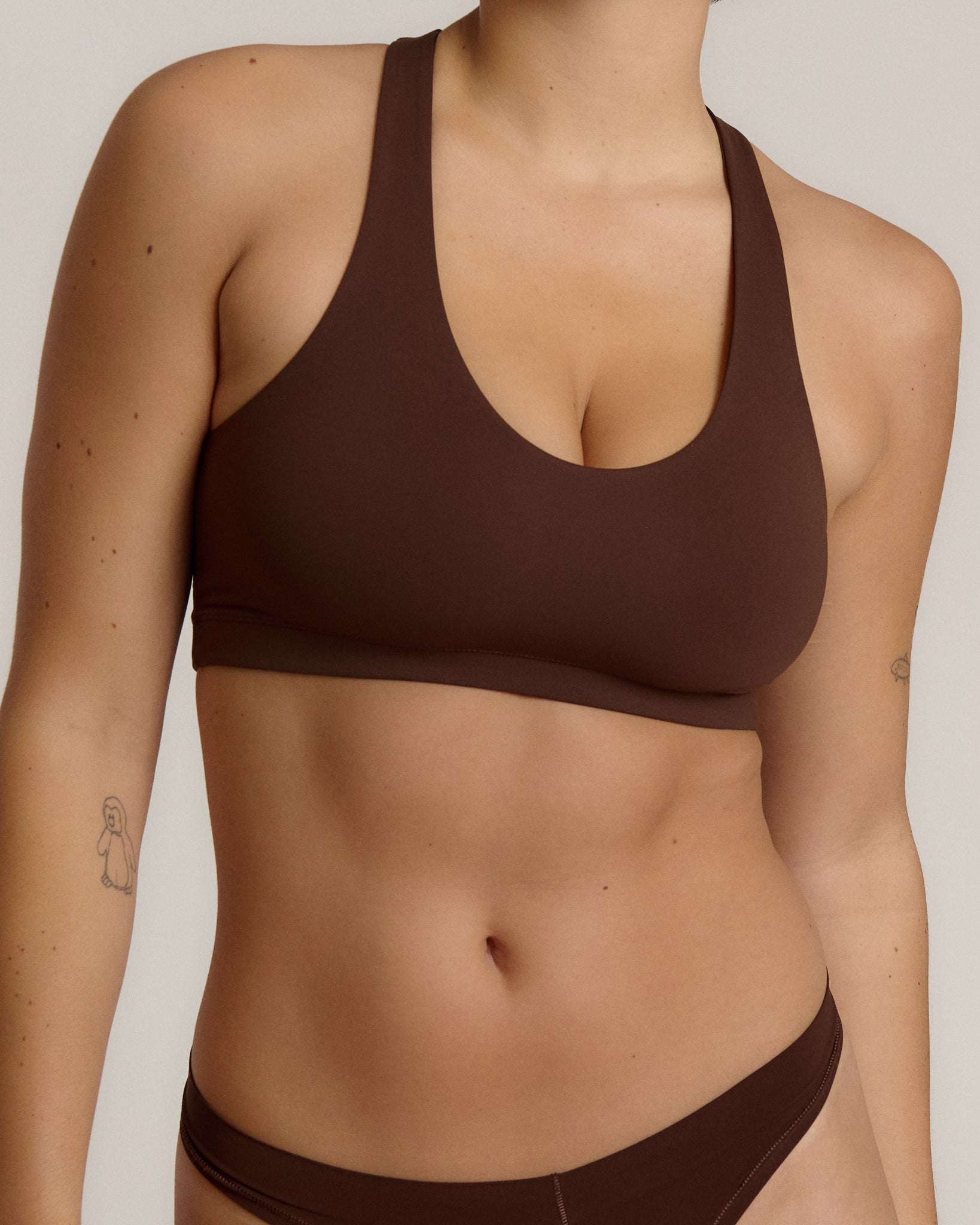 A woman modeling a matching mocha racerback and underwear set against a plain light beige studio background.