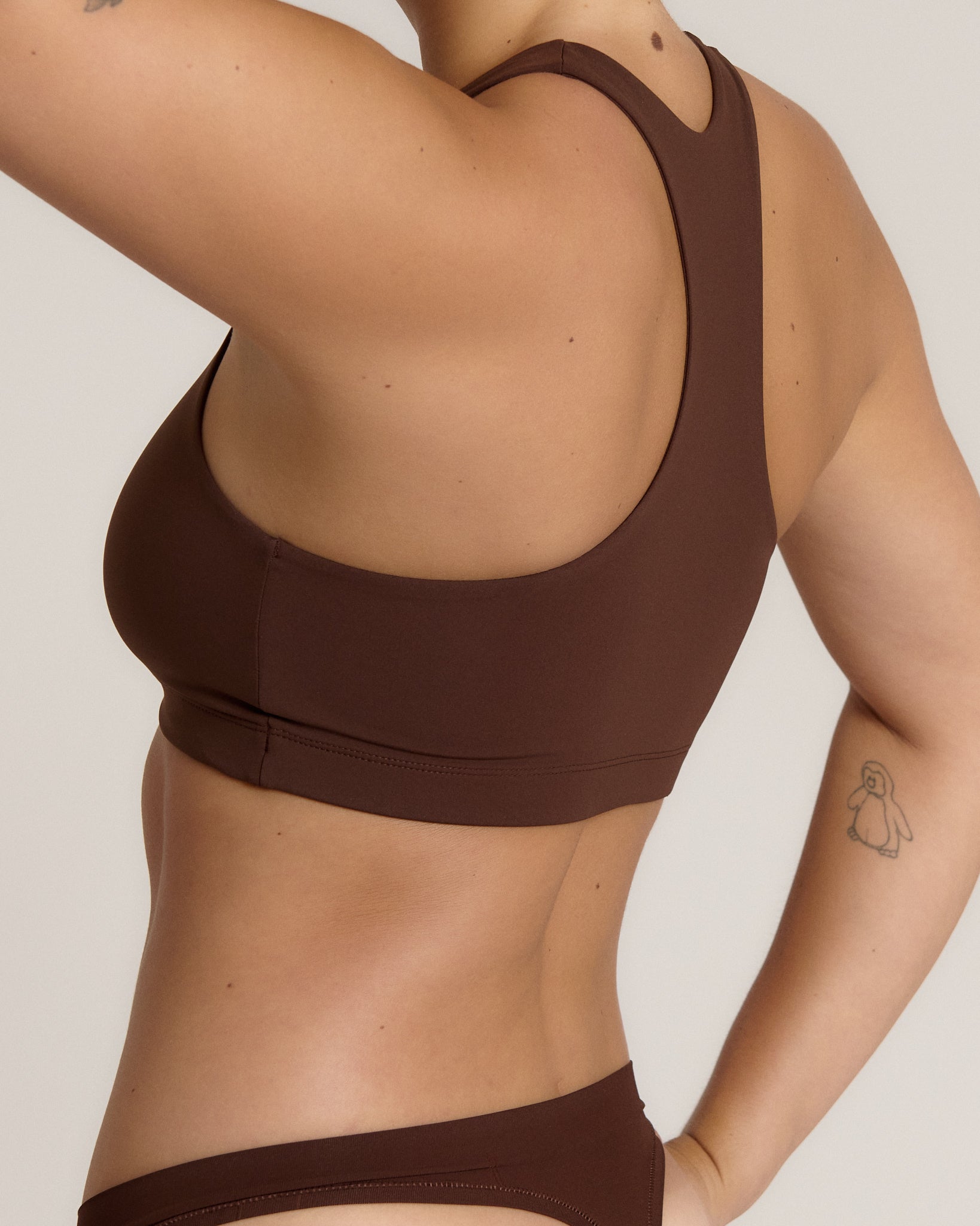A woman modeling a matching mocha racerback and underwear set against a plain light beige studio background.