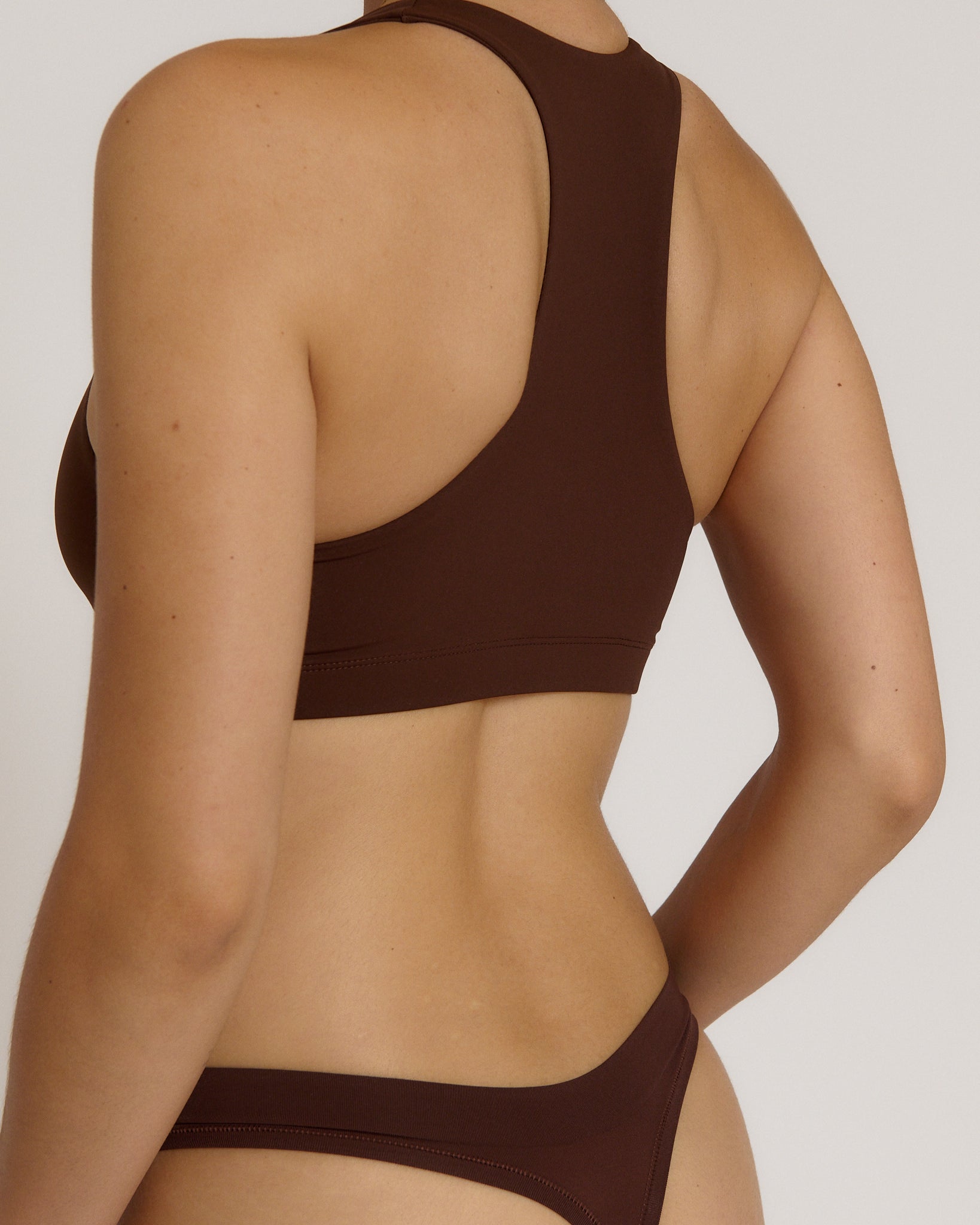 A woman modeling a matching mocha racerback and underwear set against a plain light beige studio background.