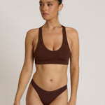 A woman modeling a matching mocha racerback and underwear set against a plain light beige studio background.