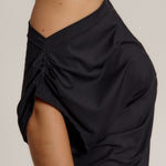 Close-up side view of a person wearing a black draped off-the-shoulder top or dress against a plain light gray background.