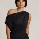 A woman modeling a black draped off-the-shoulder top against a plain light beige studio background.