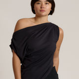 A woman modeling a black draped off-the-shoulder top against a plain light beige studio background.