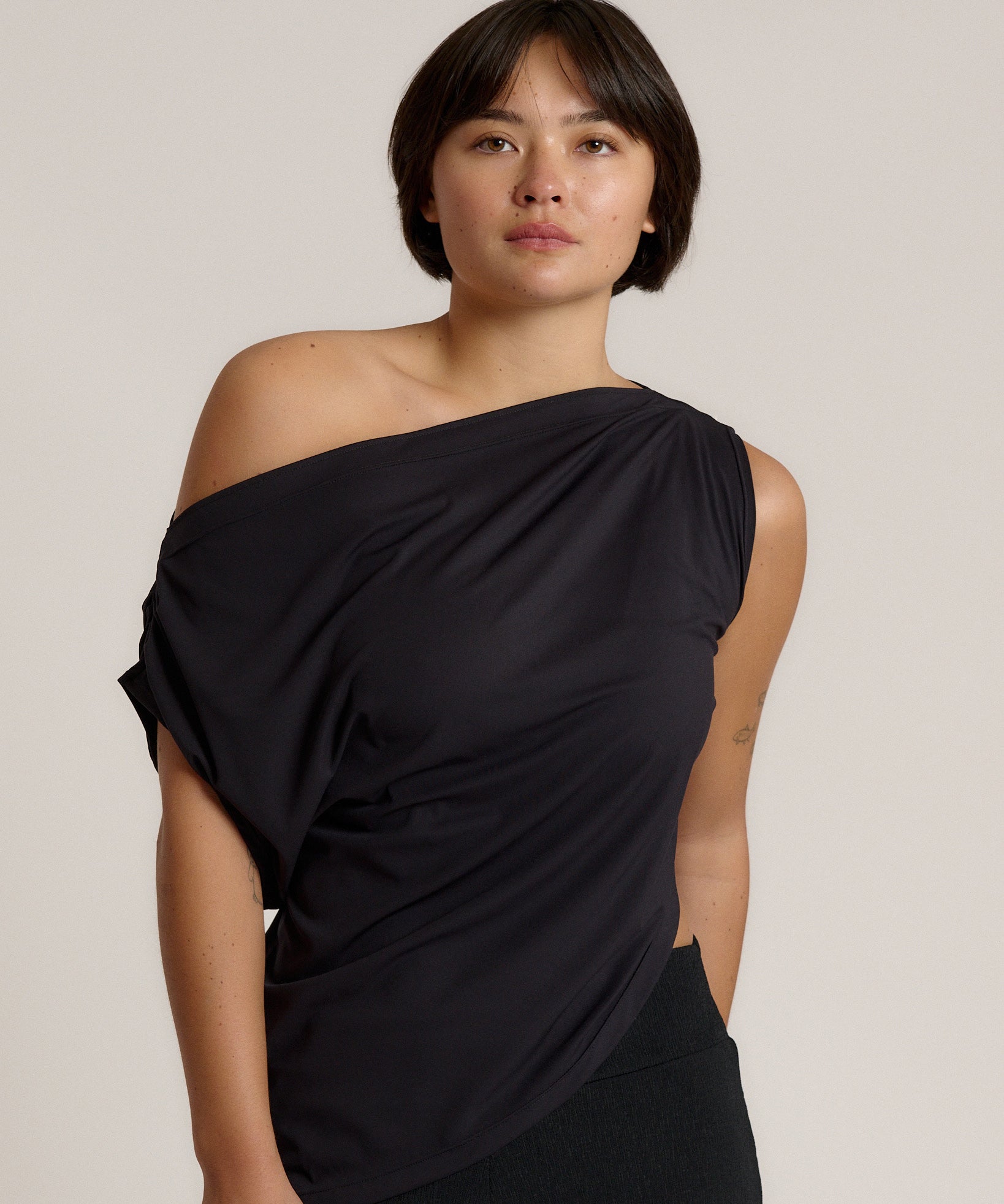A woman modeling a black draped off-the-shoulder top against a plain light beige studio background.