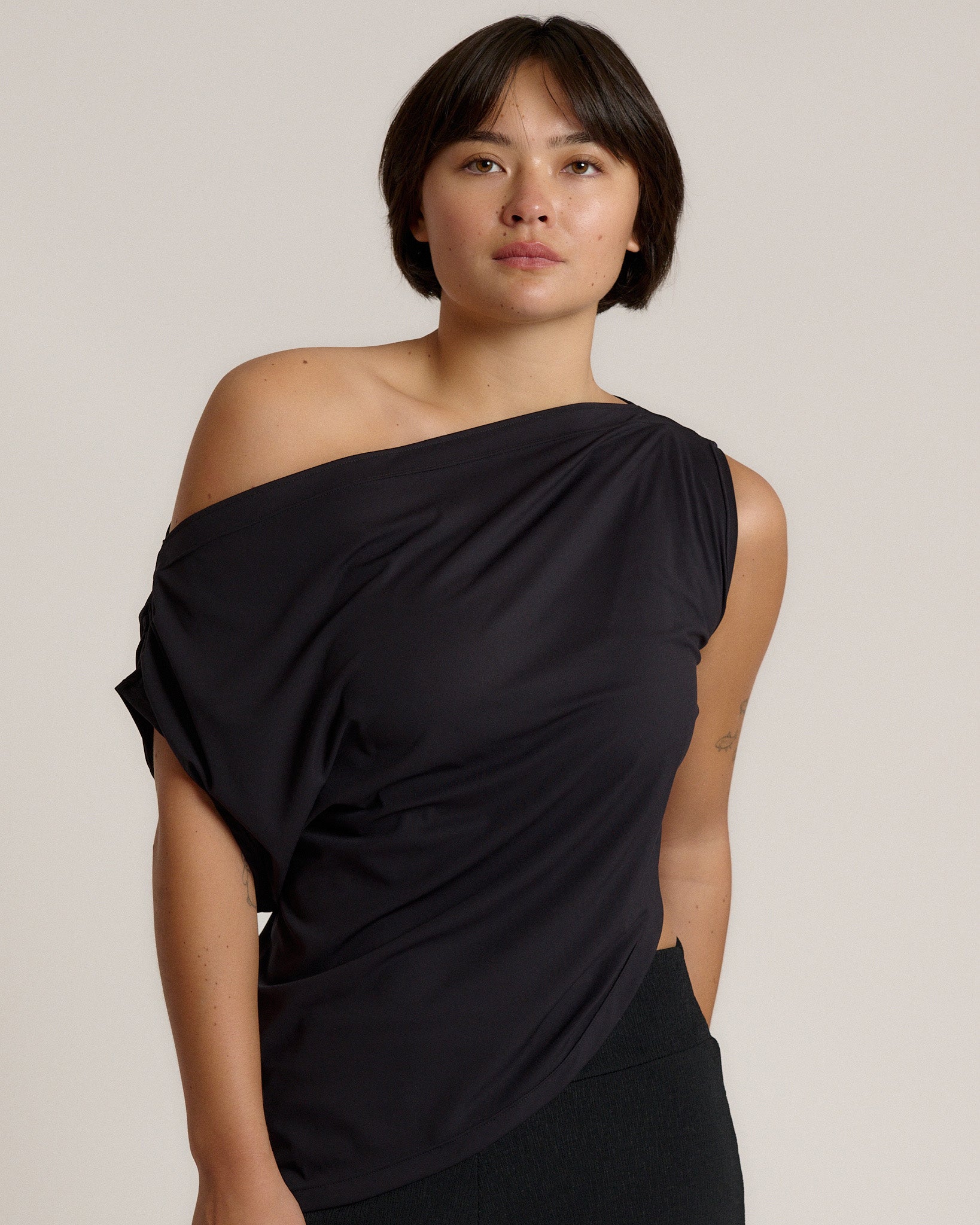 A woman modeling a black draped off-the-shoulder top against a plain light beige studio background.