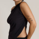 A close-up side view of a model wearing a black asymmetrical draped tank top with a side slit, set against a plain light gray background.