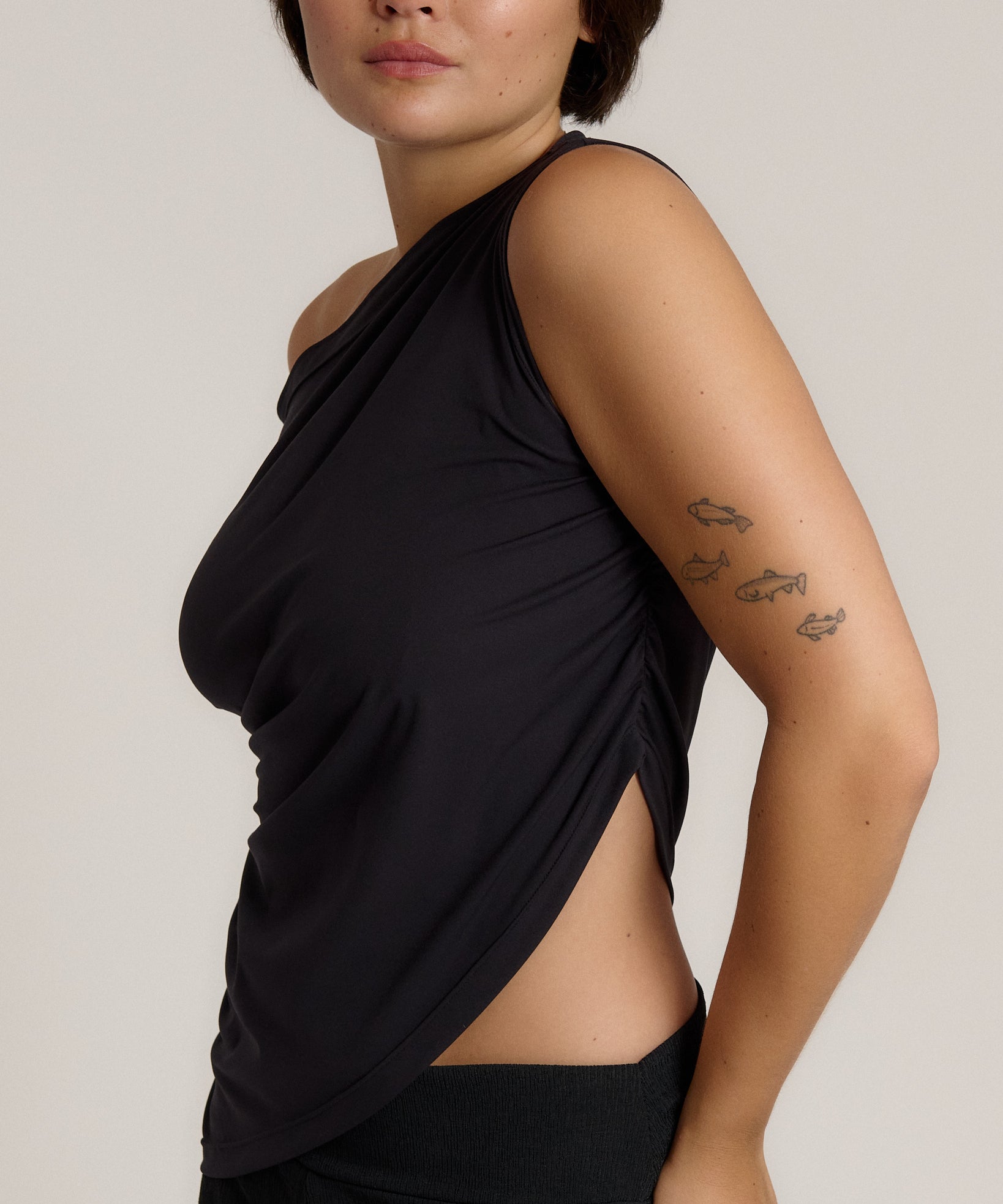 A close-up side view of a model wearing a black asymmetrical draped tank top with a side slit, set against a plain light gray background.