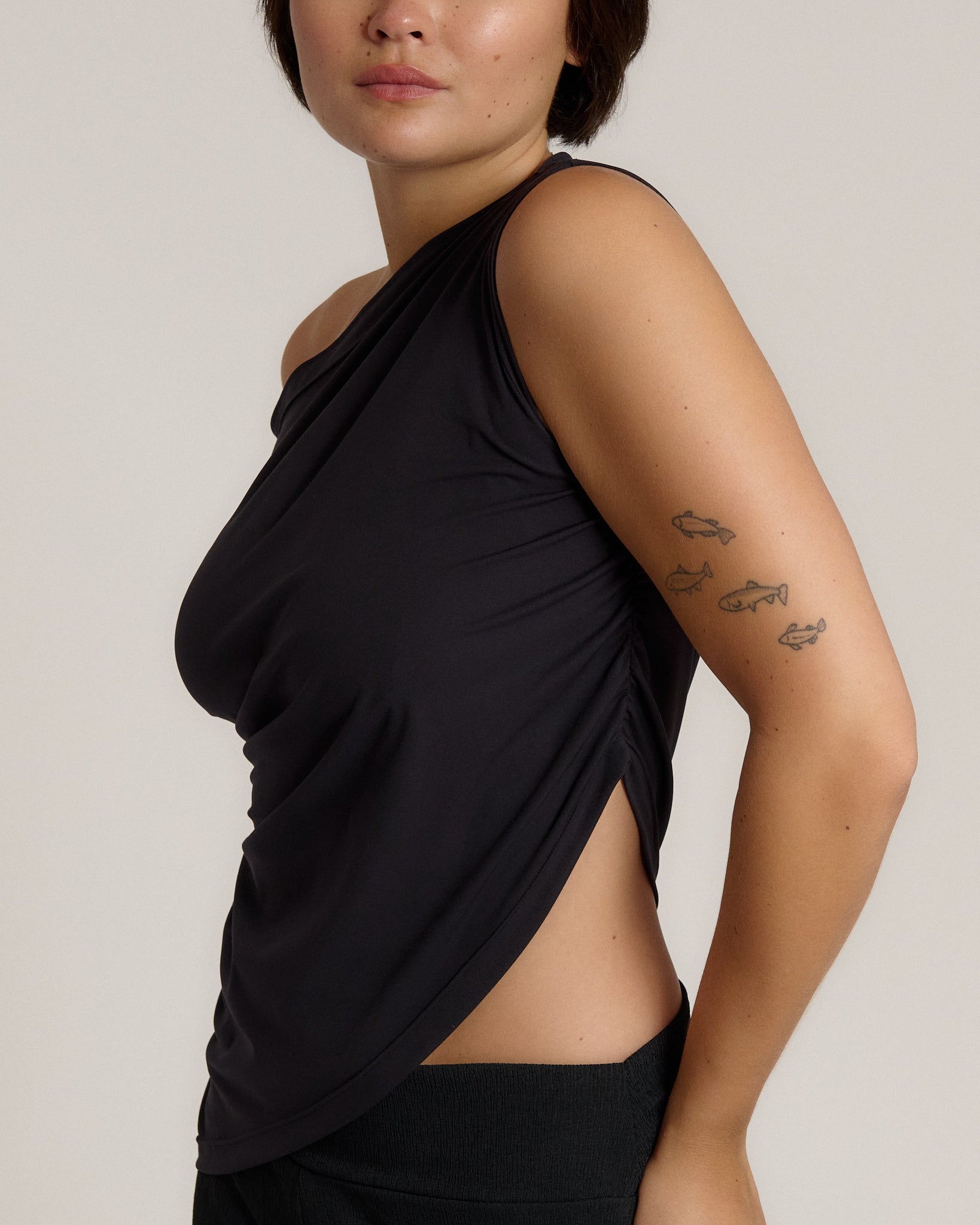 A close-up side view of a model wearing a black asymmetrical draped tank top with a side slit, set against a plain light gray background.