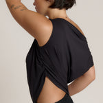A woman wearing a black draped tank top with side cutouts, shown from a side-back angle against a plain light beige studio background.
