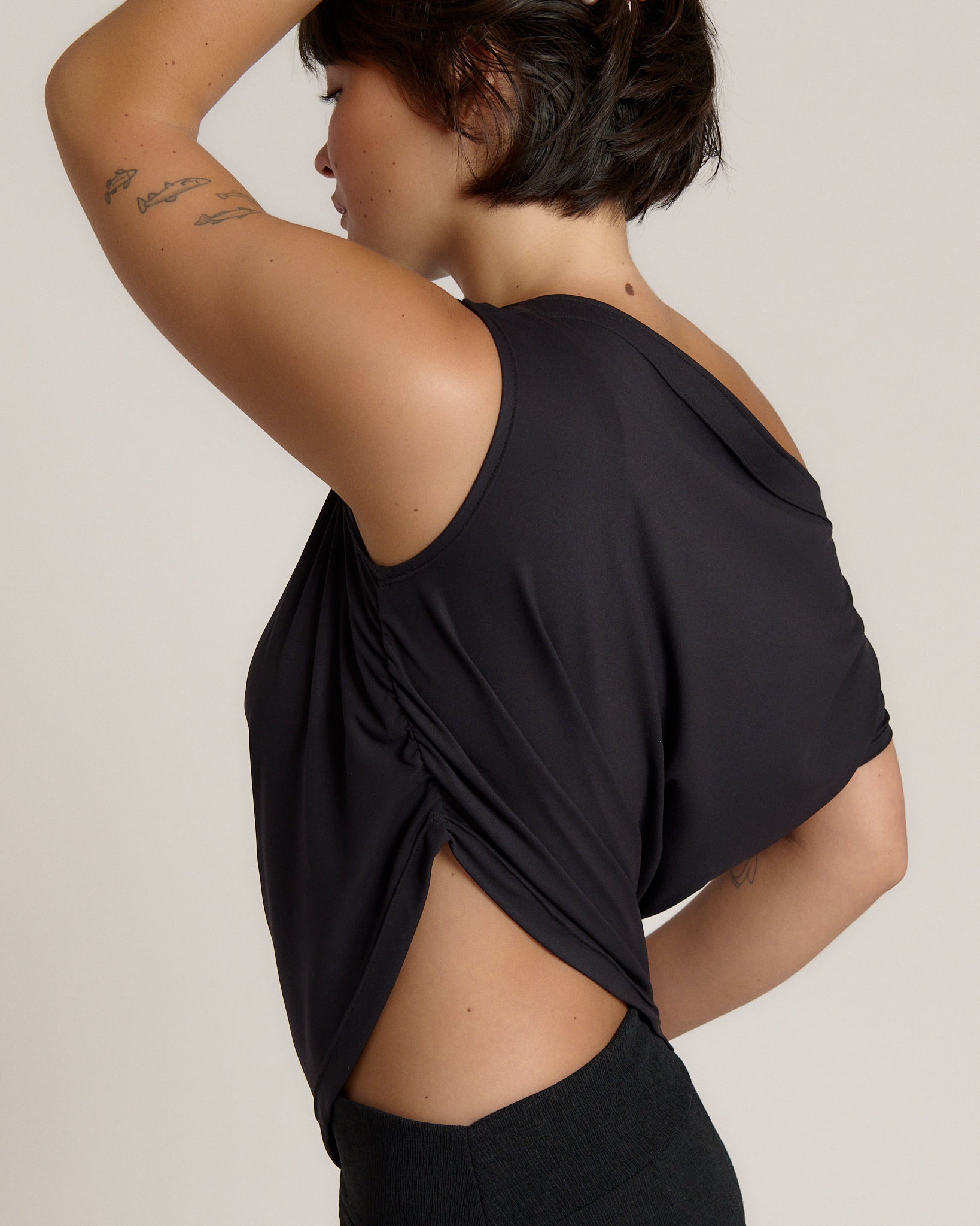 A woman wearing a black draped tank top with side cutouts, shown from a side-back angle against a plain light beige studio background.