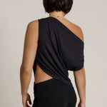 A woman shown from the back wearing a black one-shoulder draped top with a side cutout and black pants, photographed against a plain light beige studio background.