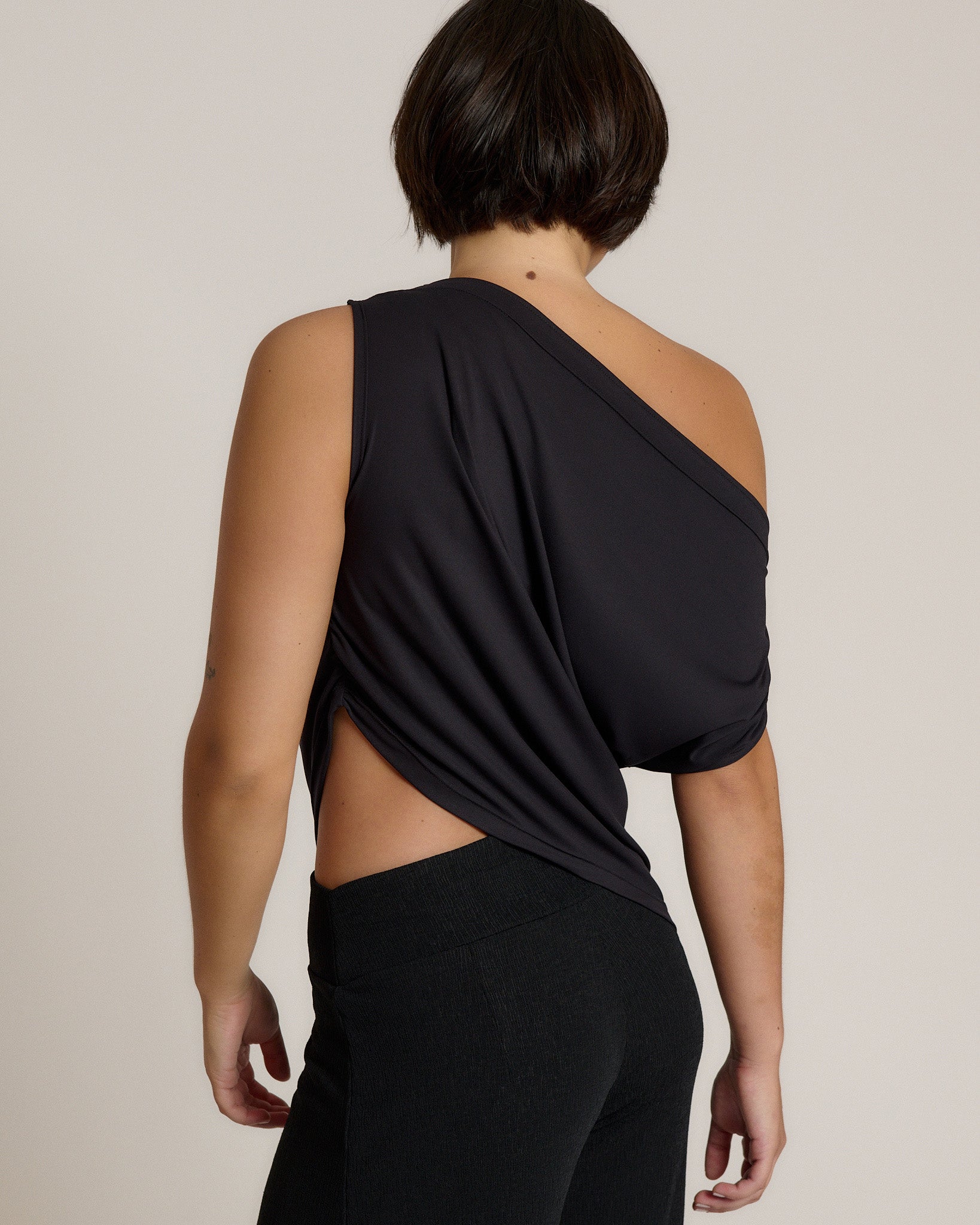 A woman shown from the back wearing a black one-shoulder draped top with a side cutout and black pants, photographed against a plain light beige studio background.
