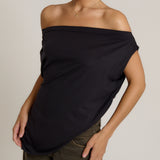 A woman wearing a black off-the-shoulder draped top and olive green pants, shown from the neck down against a light gray studio background.