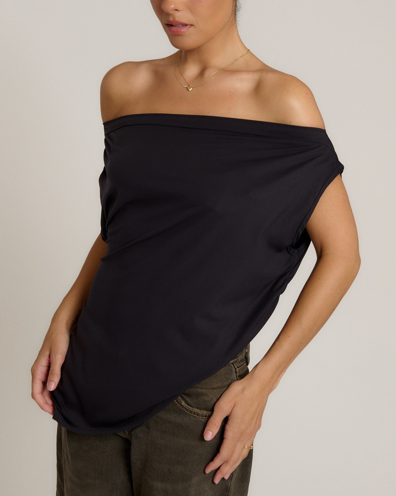 A woman wearing a black off-the-shoulder draped top and olive green pants, shown from the neck down against a light gray studio background.