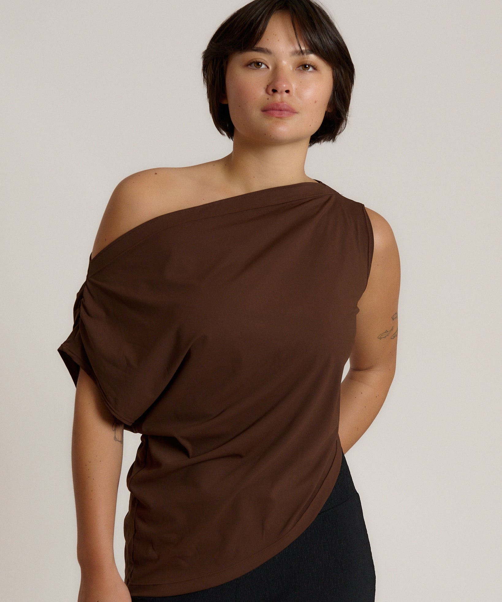 A woman modeling a draped, one-shoulder brown top against a plain light beige studio background.