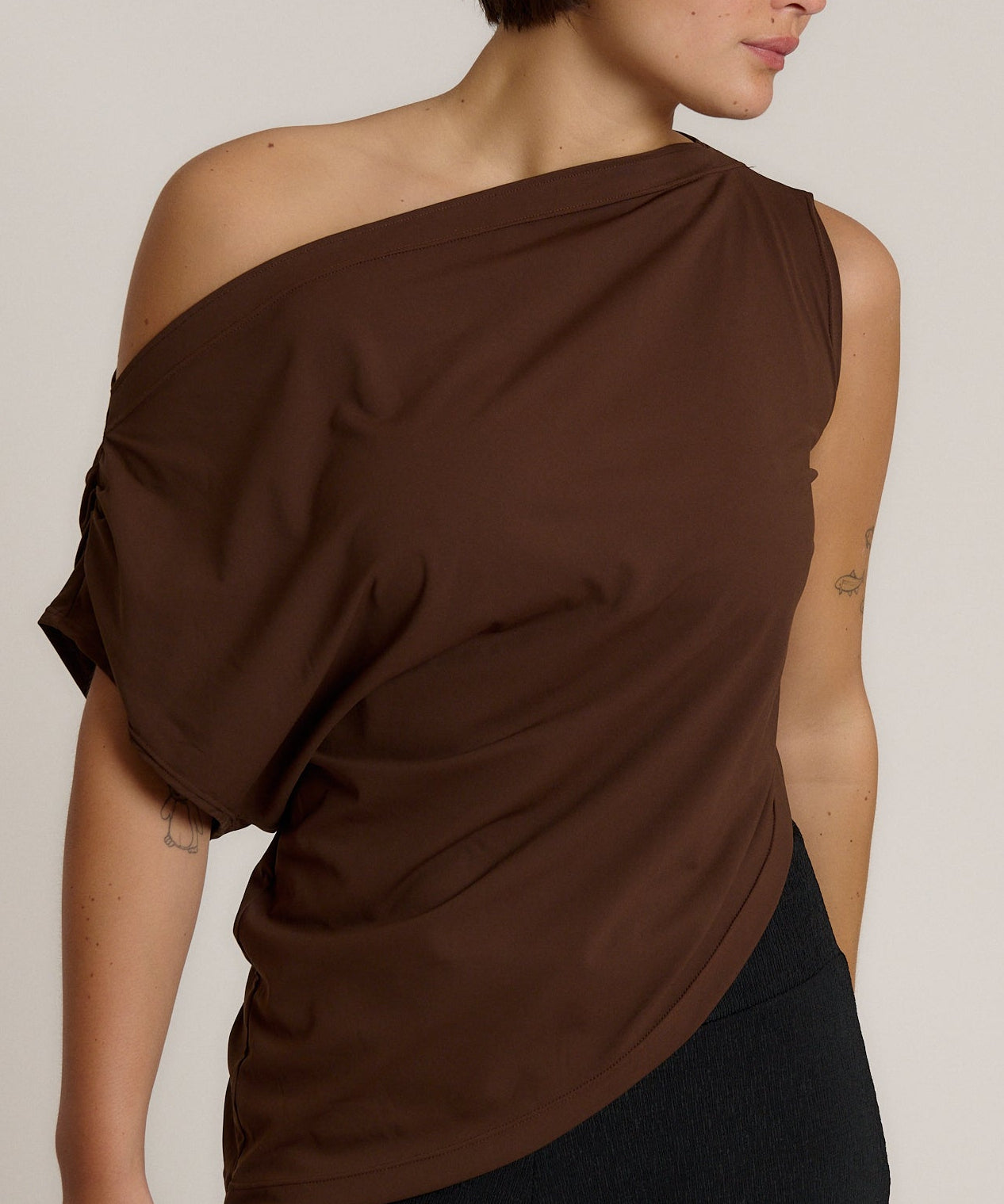 A woman wearing a dark brown off-the-shoulder draped top and black bottoms, photographed against a plain light beige studio background.
