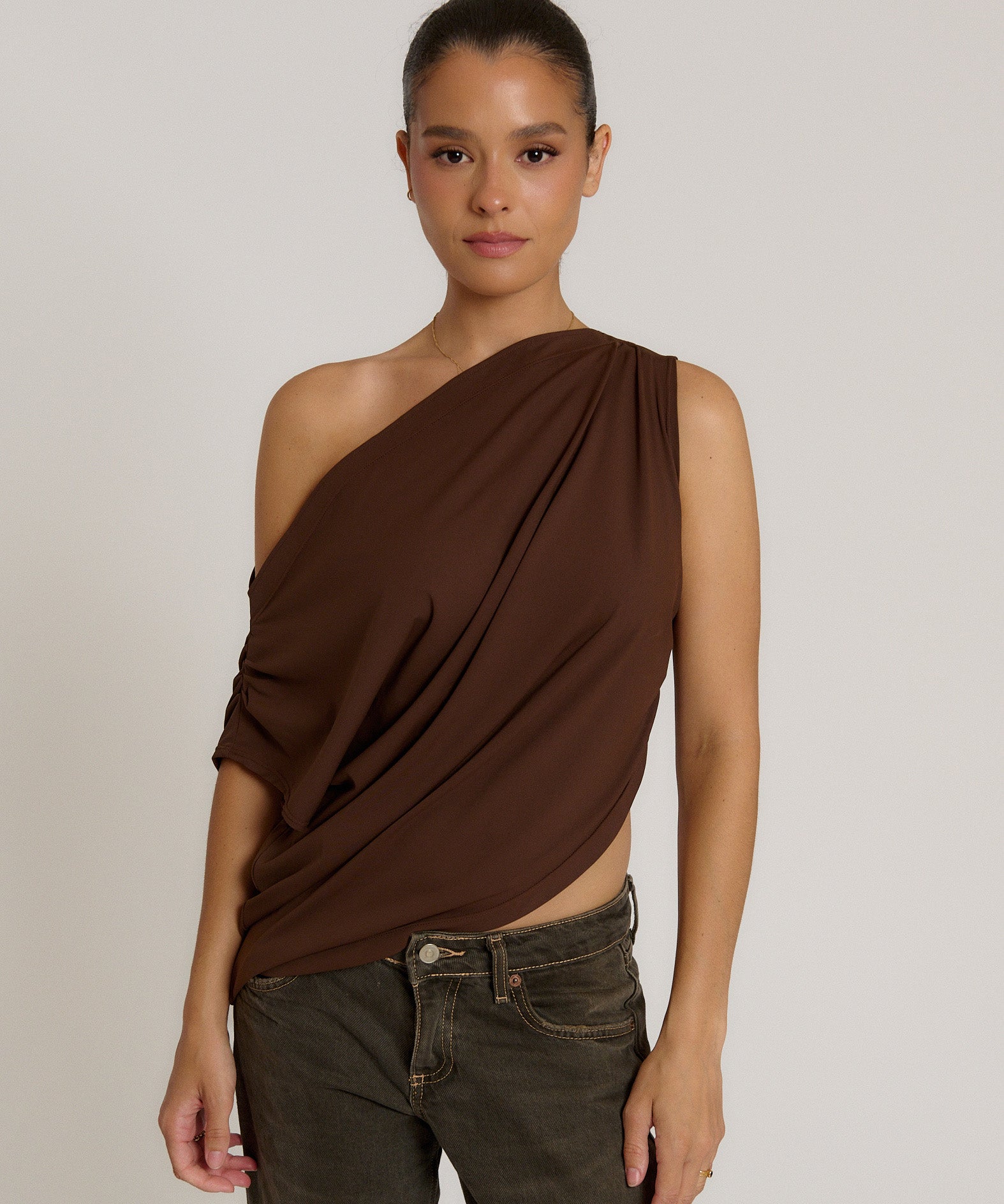 A woman modeling a draped, one-shoulder dark brown top paired with dark gray jeans, set against a plain light gray studio background.