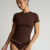 Woman wearing a brown t-shirt and underwear against a plain background