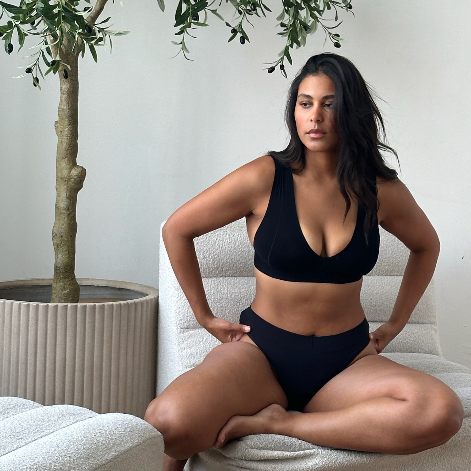 A woman wearing a black underwear set sits cross-legged on a white textured sofa next to a potted olive tree.