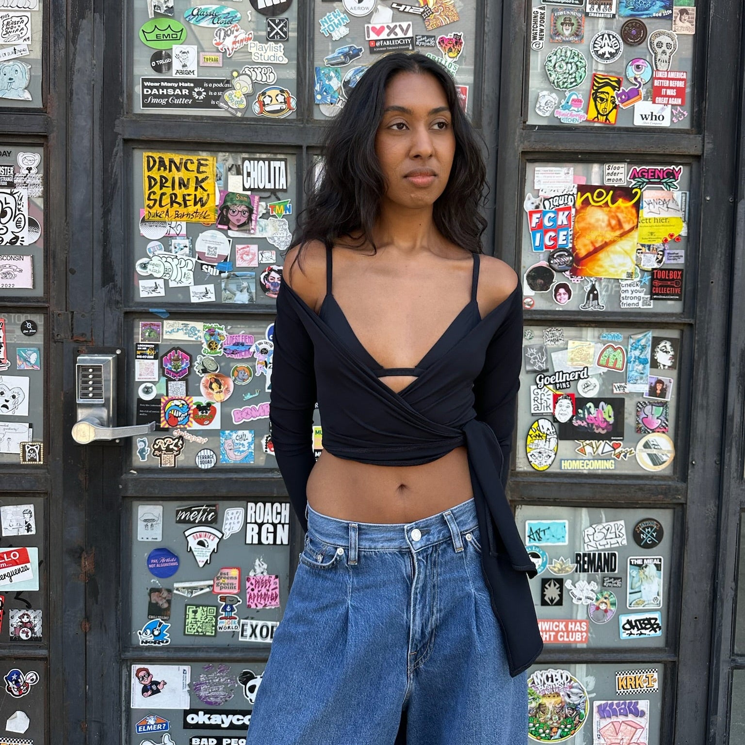 A woman stands in front of a dark door covered with colorful stickers, wearing a black off-the-shoulder wrap top and high-waisted blue jeans.