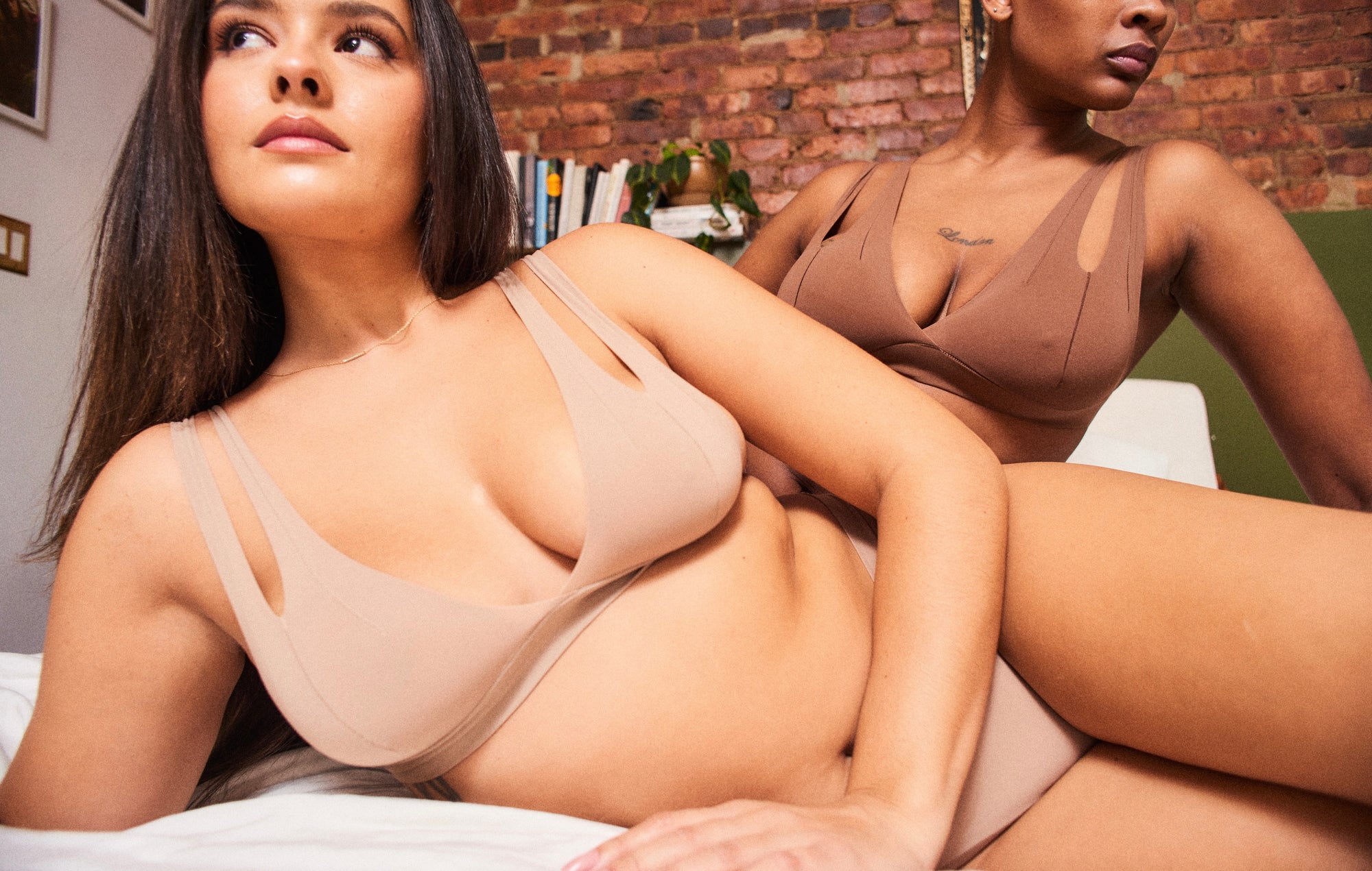 Two women laying on a bed, one wearing the Tee System and Thong in Light and the other wearing the Tee System in Brownz