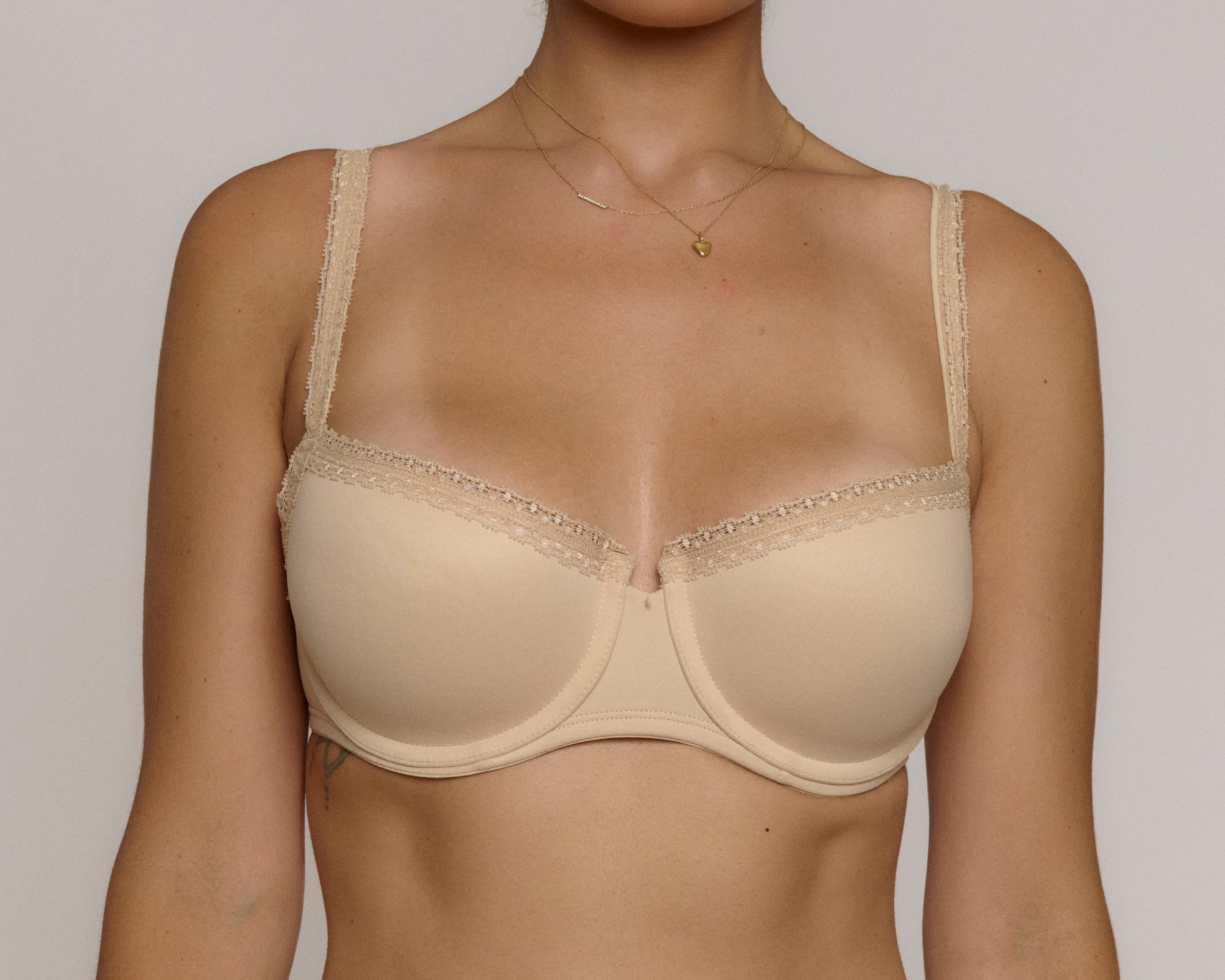 A close-up of a model wearing a beige underwire bra with lace trim along the straps and cups, set against a plain light gray background.