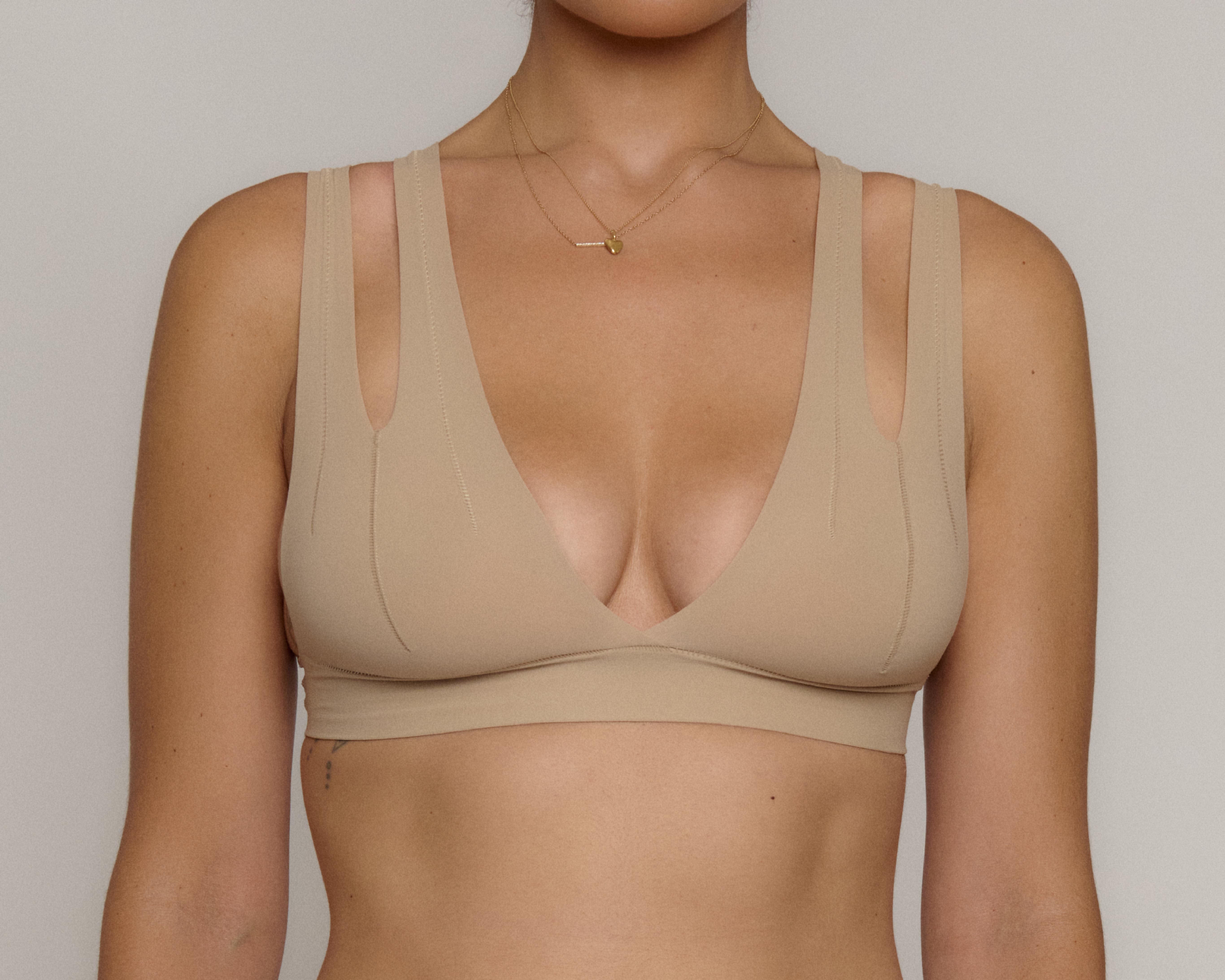 A close-up of a woman wearing a beige seamless bralette with a deep V-neckline and wide shoulder straps, shown against a neutral light gray background.